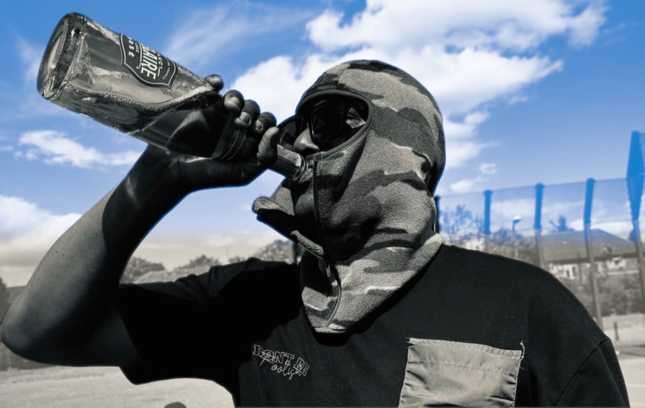 Flirta D in a camo balaclava drinking from a bottle under a blue sky