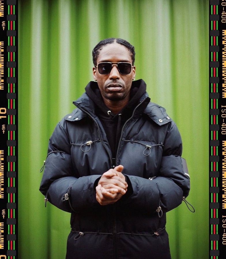 Tintz in a black puffer jacket and sunglasses against a green wall